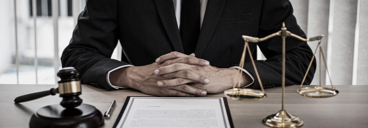 man in pak zit met gevouwen handen aan bureau. Zijn hoofd is niet zichtbaar